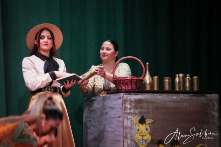 Two women in costume performing a pantomime scene with props and a green curtain background. Young actresses performing in a pantomime scene on stage.