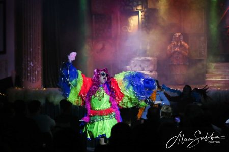 A lively scene from the pantomime "Il-Mummja" featuring a performer in a vivid, elaborate costume entertaining the audience. Colourful clown costume with feathers and bright fabric on stage.
