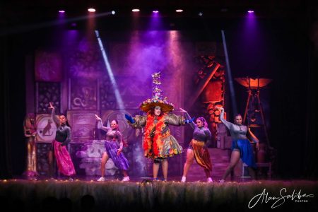 A lively scene from "Il-Mummja" pantomime featuring dancers and a central performer in elaborate costume. Vibrant pantomime performance on stage with dancers and colourful costumes.