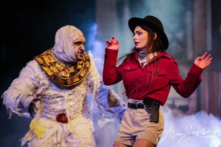 A scene from the pantomime "Il-Mummja" featuring actors in costume during a theatrical performance. An actor in a mummy costume and a woman in a red jacket and hat perform on stage.