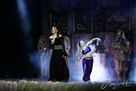 Performers in vibrant costumes perform a scene from "Il-Mummja" pantomime on stage with atmospheric lighting. Elegant performers in traditional costumes during a pantomime show.