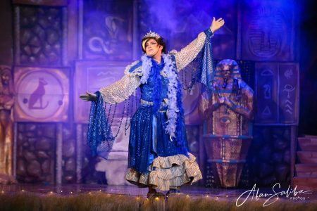 A performer dressed in vibrant costume performing in a pantomime titled "Il-Mummja". A performer in colourful costume on stage during a pantomime show.