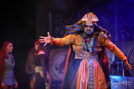 A theatrical scene from "Il-Mummja" featuring a performer in vibrant costume with expressive gestures. Performer in colourful costume acting in a pantomime scene.