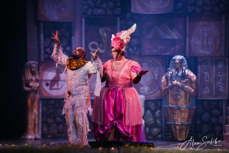 Performers in colourful costumes during a theatrical pantomime show. An actress in a vibrant pink costume with a large feathered headpiece performs on stage.