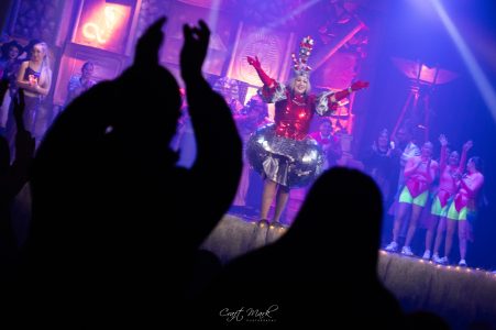 A performer in colourful costume on stage during a pantomime show, with an audience clapping and enjoying the performance. Pantomima - Il-Mummja performance at Fondazzjoni Teatrali.