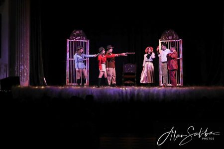 A theatrical scene from "Il-Mummja" featuring actors in period costumes with props and set pieces. Performers in costume during a pantomime scene on stage.
