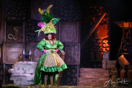 A performer dressed in a vibrant costume with a large feathered hat during a theatrical performance of "Pantomima - Il-Mummja". Vibrant theatre scene featuring a performer in a colourful costume with an elaborate feathered hat.