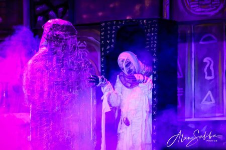 A scene from the pantomime "Il-Mummja" featuring performers in elaborate costumes and atmospheric purple lighting. Performers in costume during a pantomime scene with mystical lighting.