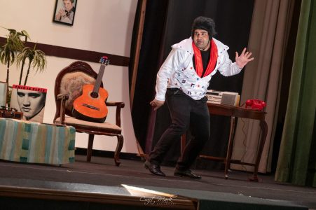 A comedian in a white jacket and black pants performs on stage with props and vintage furniture. Performer in a comedic act on stage with a guitar and vintage decor.