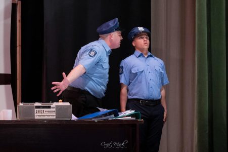 Two actors dressed as police officers performing a scene on stage, with a desk and props in the background. Police officers on stage during a theatrical performance.