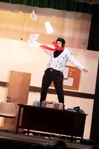 A theatrical performance by Fondazzjoni Teatrali Silvio Fleri Soler, showcasing an actor in a classroom scene with papers flying around. Performer on stage with papers flying, in a classroom setting.