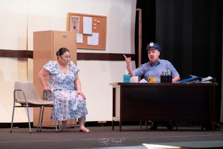 A scene from a theatrical performance featuring two actors, one seated and one standing, in a classroom setting. Actors performing on stage in a theatre production.
