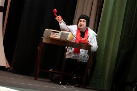 Actor in a theatrical performance, using a vintage telephone and desk, showcasing the foundation's focus on theatrical arts. Comedian performing on stage with vintage telephone and desk.