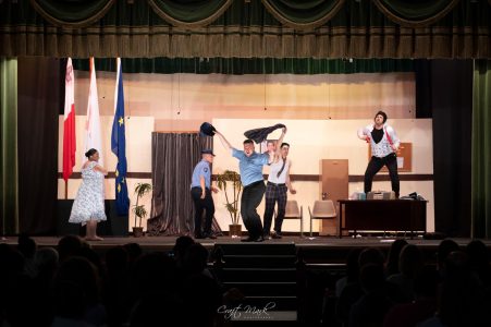 A group of actors performing a play on stage, with a backdrop and audience watching attentively. Performing a theatrical scene on stage with actors and props.