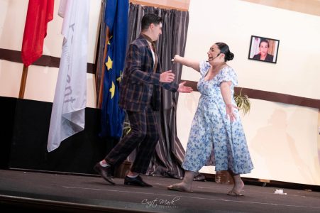 A scene from a theatrical play featuring two actors engaged in an intense moment, with flags and a screen in the background. Actors performing a theatrical scene on stage during a live performance.