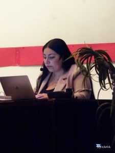 A woman focused on her laptop at a comedy night event, with a potted plant nearby and a simple background. Woman working on a laptop during a comedy night event.