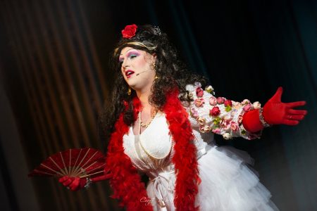 A performer dressed in vibrant theatrical costume with fan and red feather boa on stage during a performance. Performer in colourful theatrical costume with fan and red feather boa.