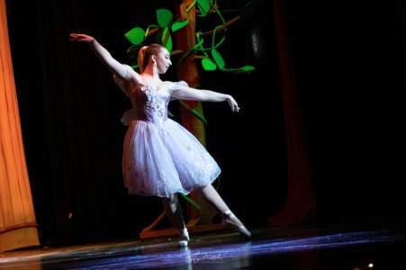 A young ballet dancer in a white tutu performs a graceful move during a stage performance. Ballet dancer performing on stage with a tree backdrop at the theatre.