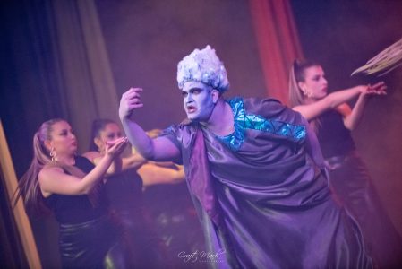 A theatrical scene featuring a performer with elaborate makeup and costume, surrounded by dancers on stage. A performer in a theatrical production with dancers in the background.