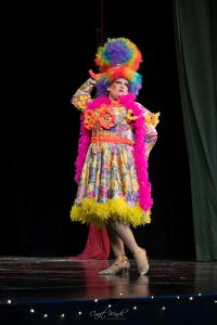 A performer in a bright, elaborate costume with feathers and floral accents on stage. Colorful theatrical costume with feathers and floral details.