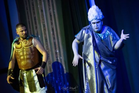 A theatrical scene featuring two actors in colorful costumes, one with a white wig, performing passionately. Actors in vibrant costumes perform a theatrical scene on stage.