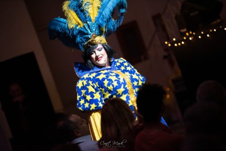 A performer dressed in a colourful star-themed costume with a large feathered headdress, entertaining an audience at a theatrical event. Performer in vibrant blue and yellow star costume with feathered headdress during a theatrical event.