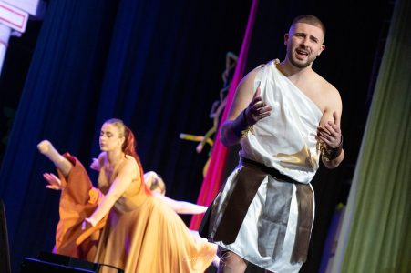 A male actor in ancient costume delivering lines during a stage performance, with female actors in the background. Actor performing in a theatrical play at Fondazzjoni Teatrali Silvio Fleri Soler.