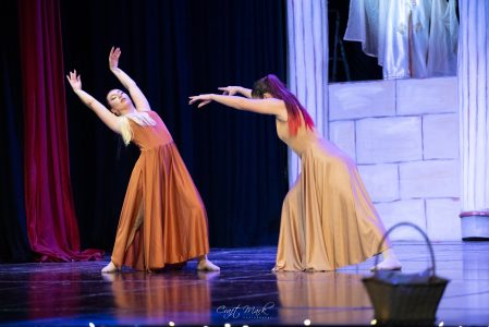 Two dancers in flowing costumes perform an expressive dance on stage with a dark backdrop. Performers dancing gracefully on stage during a theatrical event.