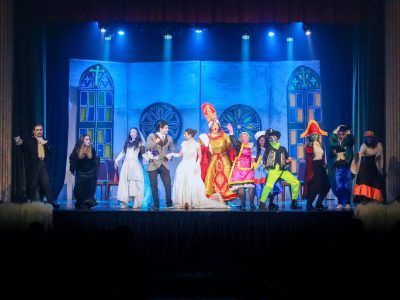 The cast of Korps Brajd performing a lively pantomime on stage with vibrant costumes and theatrical backdrop. Cast performing in a colourful pantomime on stage with set design.