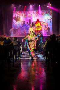 Vivid costumes and a lively performance entertain an audience in a theatrical setting. Brightly coloured performers on stage during a pantomime show.